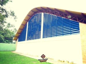 Veneziana para Galpão A imagem mostra a veneziana industrial Solution com pintura eletrostática Acul del Rey instalada na fachada de um ginásio poliesportivo em formato de arco. Essa aplicação tem como finalidade principal proporcionar um sistema eficiente de ventilação e controle de iluminação natural para o interior do ginásio, além de agregar um aspecto estético marcante.A veneziana industrial Solution é especialmente projetada para permitir a circulação de ar adequada no espaço, auxiliando na dissipação de calor, controle de umidade e renovação do ar. Isso é essencial em ambientes como ginásios poliesportivos, que costumam ser frequentados por um grande número de pessoas, gerando calor e uma demanda maior por conforto térmico. Além disso, a instalação da veneziana em formato de arco na fachada do ginásio proporciona um visual impactante e diferenciado, conferindo personalidade e identidade ao local. A pintura eletrostática Acul del Rey, por sua vez, não só contribui para a estética da fachada, dando um toque de elegância, mas também oferece proteção contra corrosão e intempéries, conferindo durabilidade à estrutura. Com essa combinação de funcionalidade e apelo visual, a veneziana industrial Solution com pintura eletrostática Acul del Rey instalada na fachada de um ginásio poliesportivo em formato de arco se torna uma solução eficiente, segura e esteticamente agradável para garantir o conforto e a qualidade do ambiente esportivo.