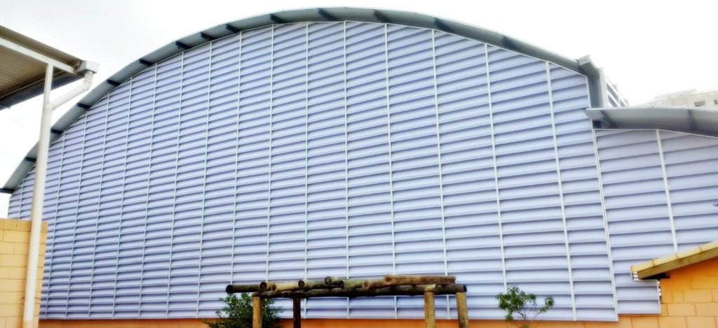 Veneziana Industrial em PVC Finalidade da Imagem:** A imagem das venezianas industriais "Solution" em policarbonato alveolar branco leitoso, instaladas em arco em um ginásio poliesportivo, busca atender aos seguintes propósitos: 1. **Destaque para Inovação:** Mostrando o produto aplicado em uma estrutura em arco, a imagem enfatiza a versatilidade e adaptabilidade das venezianas industriais, sugerindo que elas não se limitam apenas a instalações padrão. 2. **Realce de Uso Específico:** A colocação em um ginásio poliesportivo indica aos clientes potenciais que o produto é adequado para grandes estruturas esportivas, garantindo iluminação e ventilação adequadas enquanto mantém uma estética moderna. 3. **Apresentação da Estética:** O policarbonato alveolar branco leitoso dá uma sensação etérea e sofisticada, indicando que o produto não é apenas funcional, mas também esteticamente agradável, perfeito para espaços que querem combinar forma e função. 4. **Demonstração de Aplicação em Grande Escala:** Ao ser instalado em um ginásio, a imagem mostra que o produto pode ser usado em projetos de grande escala, atendendo às necessidades de instalações maiores. 5. **Promoção e Engajamento:** Uma instalação impressionante como essa pode atrair mais atenção nas campanhas de marketing, funcionando como uma peça central em materiais promocionais, seja online ou offline. 6. **Prova Social:** Mostrar o produto em uso em um ambiente tão público e amplamente utilizado como um ginásio poliesportivo fornece uma forma de prova social, indicando que instituições confiáveis optam por esse produto para suas necessidades.