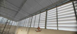 Veneziana Industrial Aleta em Fibra Aleta em Fibra de Vidro A imagem apresenta um galpão industrial espaçoso e bem iluminado, com várias venezianas da Solution Venezianas Industriais instaladas nas laterais. As venezianas estão parcialmente abertas, permitindo a entrada de luz natural e ar fresco. Trabalhadores são vistos no chão do galpão, operando máquinas e equipamentos, claramente beneficiados pela excelente ventilação e iluminação. O logotipo da Solution Venezianas Industriais é visível no canto inferior direito.