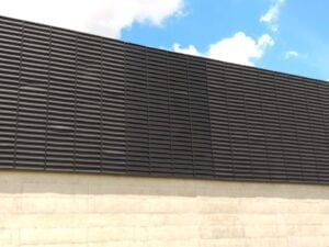 Veneziana Industrial na Fachada Veneziana Industrial para Fachada Industrial, A imagem exibe uma fachada industrial onde uma "Veneziana Industrial" da Solução está instalada. As venezianas têm lâminas planejadas que estão posicionadas de forma a permitir a entrada controlada de luz natural. Algumas lâminas estão abertas, permitindo que a luz do dia ilumine o interior da instalação, enquanto outras estão fechadas para bloquear o excesso de luz. Isso demonstra o controle preciso que as venezianas oferecem sobre a iluminação. A fachada em si apresenta um design moderno que se integra harmoniosamente com o ambiente industrial circundante. A imagem transmite a ideia de eficiência energética, conforto térmico e uma estética atraente, características fundamentais das indústrias venezianas da Solution.