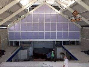 Solution Venezianas Industriais, A imagem mostra uma veneziana industrial instalada em um ambiente de fábrica. A principal finalidade dessa veneziana é melhorar a ventilação e a iluminação natural do espaço, garantindo um ambiente de trabalho mais saudável e eficiente. Feita de materiais duráveis, como alumínio ou policarbonato, a veneziana industrial é ideal para instalações que exigem robustez e resistência às condições ambientais. Seu design funcional permite a circulação de ar fresco e a entrada de luz, reduzindo a necessidade de iluminação artificial e melhorando a qualidade do ar dentro do ambiente industrial.